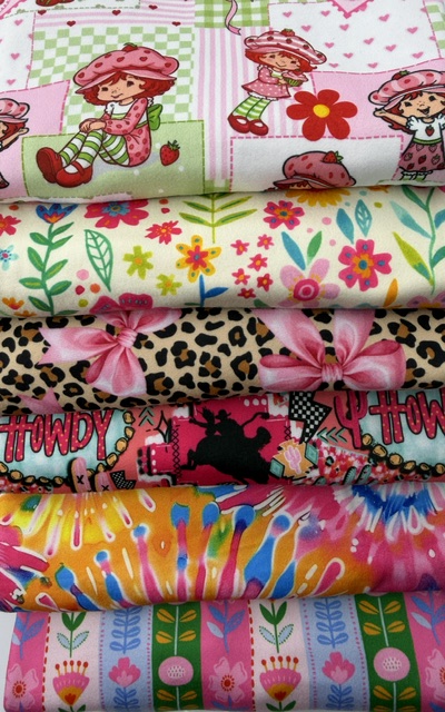 Stack of folded double brushed poly knit fabric bolts in strawberry girl, retro floral, pink leopard bow, western howdy, tie-dye and folk flower stripe prints